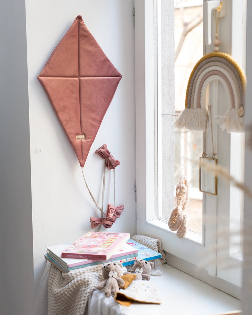 A cozy, well-lit corner by a window features the Oeko-Tex®Standard100 certified Velvet Kite Wall Decor in Dusty Pink with matching bows, a rainbow wall hanging, and a small stack of children's books on a table. This charming setup is completed with crocheted toys and a soft knitted blanket—perfect for children's room decoration.