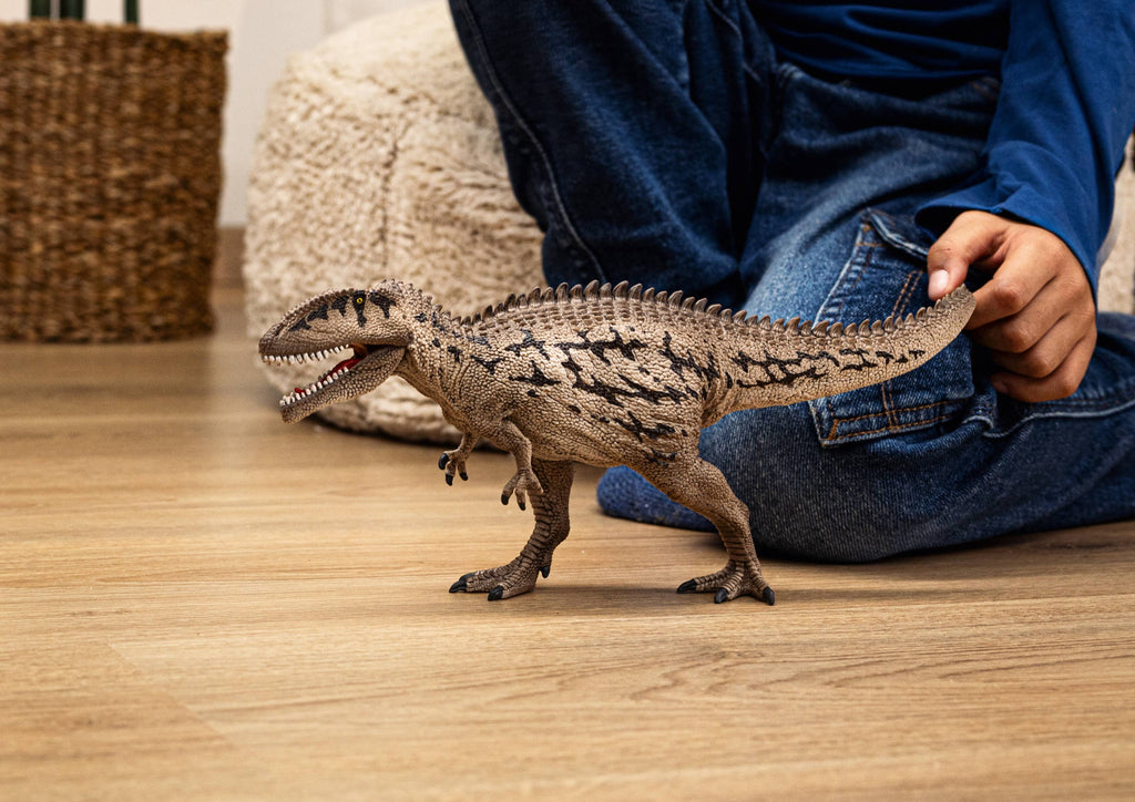 A child in blue jeans and a blue shirt sits on a wooden floor, holding the Schleich Carcharodontosaurus Dinosaur Toy by its tail. The large, detailed dinosaur figure features a movable jaw. A woven basket and a cream pouf are visible in the background.