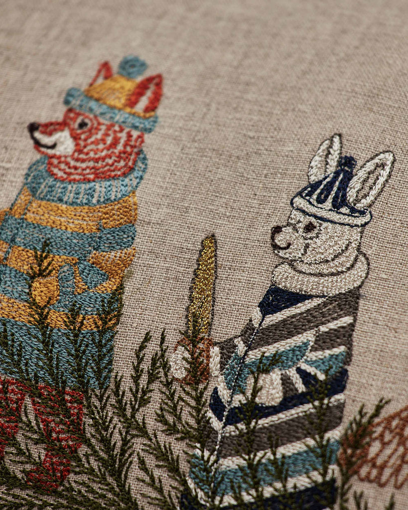 The Coral & Tusk Christmas Tree Sled Pillow features embroidered fox and rabbit friends in festive sweaters and hats, surrounded by green branches on beige fabric—a charming holiday accent for your home décor.