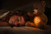 A child sleeps peacefully under a blanket with the Light at Night Caspar Teddy Bear Plush, emitting a soft warm glow on her face. The bear's built-in USB charger enhances the cozy atmosphere in the dimly lit room.