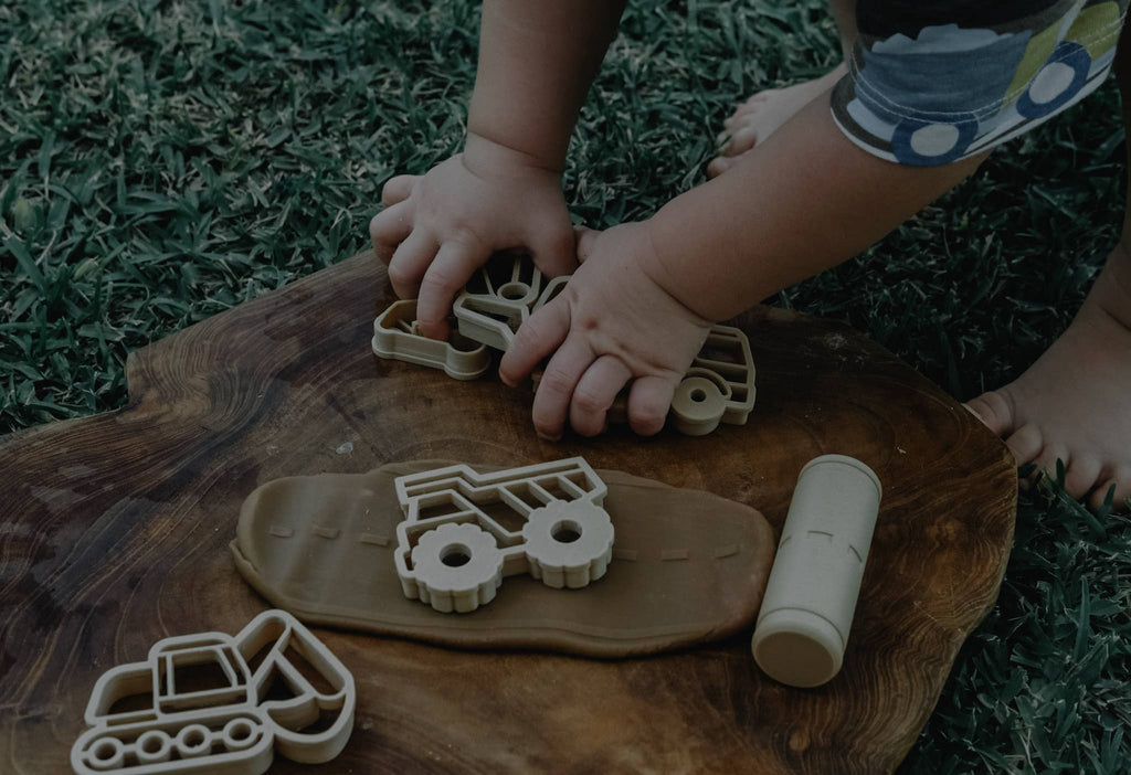 A child enjoys sensory play outdoors, using the Construction Cutter Set - Eco Playdough Tool to shape dough into construction vehicles on a wooden board, with grass in the background.