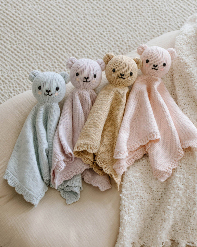 A soft, hand-knit Cuddle + Kind Organic Cotton Baby Bear Lovey in Baby Blue rests on a cozy cream surface. This adorable lovey features an organic cotton bear head and blanket body, perfect for your little one to cherish.