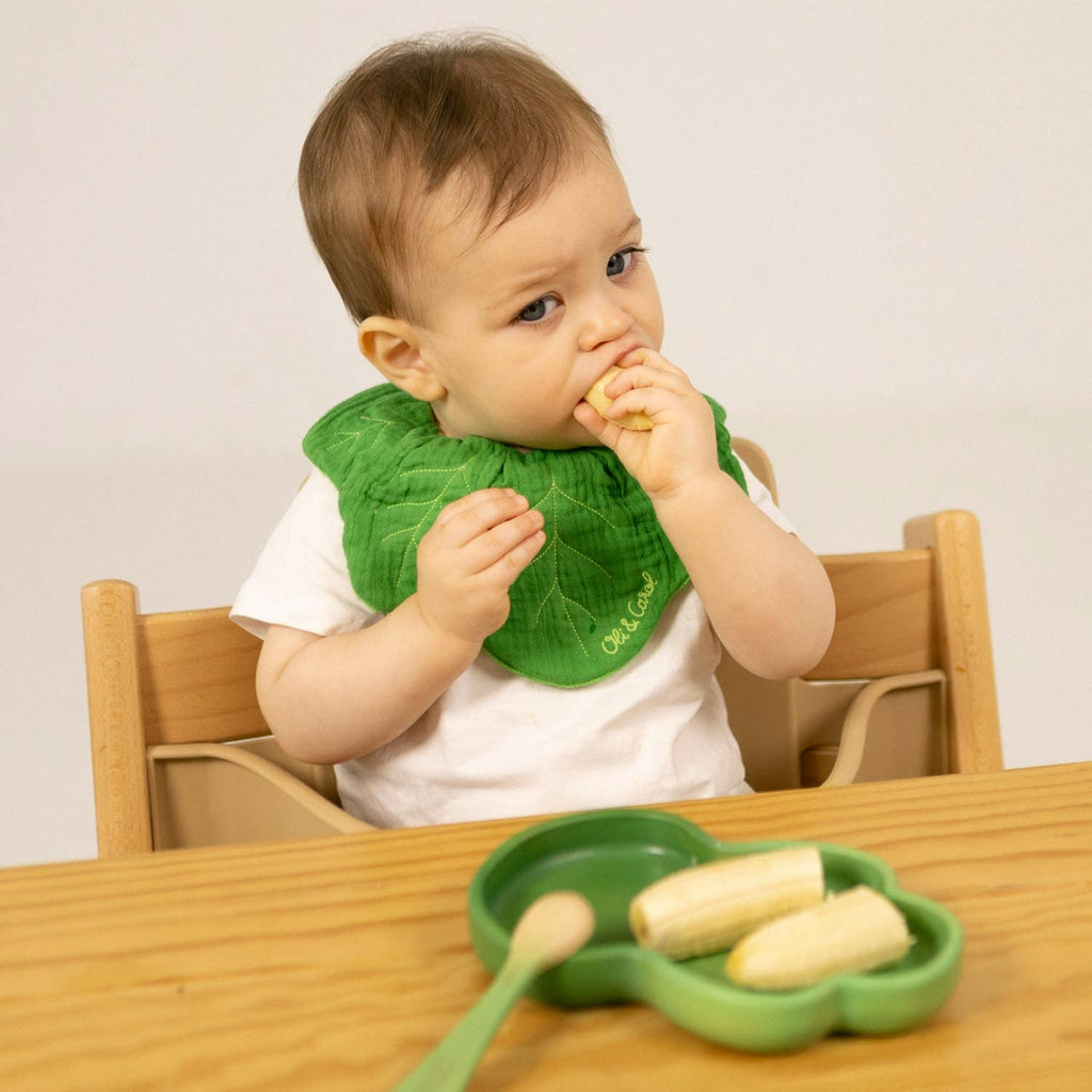 A baby wearing a green bib sits at a wooden table, eating a piece of banana from the Kendall the Kale Plate & Spoon Set, which features a plate shaped like a clover.