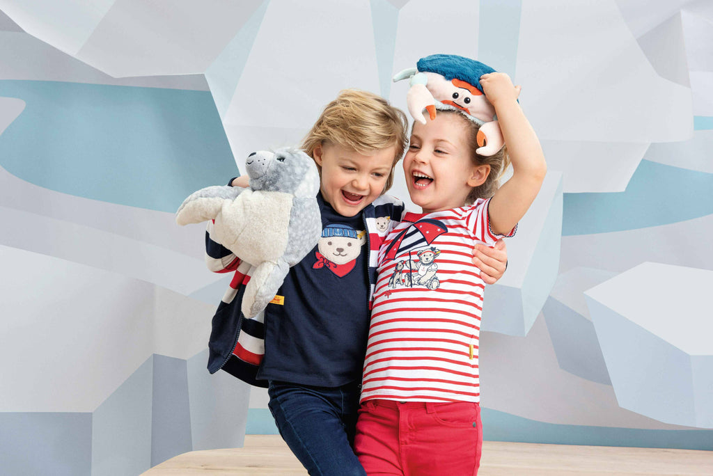 Two smiling children hug and laugh. One holds a Steiff Curby Crab Plush Stuffed Toy, 9 Inches, above their head; both wear colorful outfits with playful prints, standing before a blue and white geometric background.