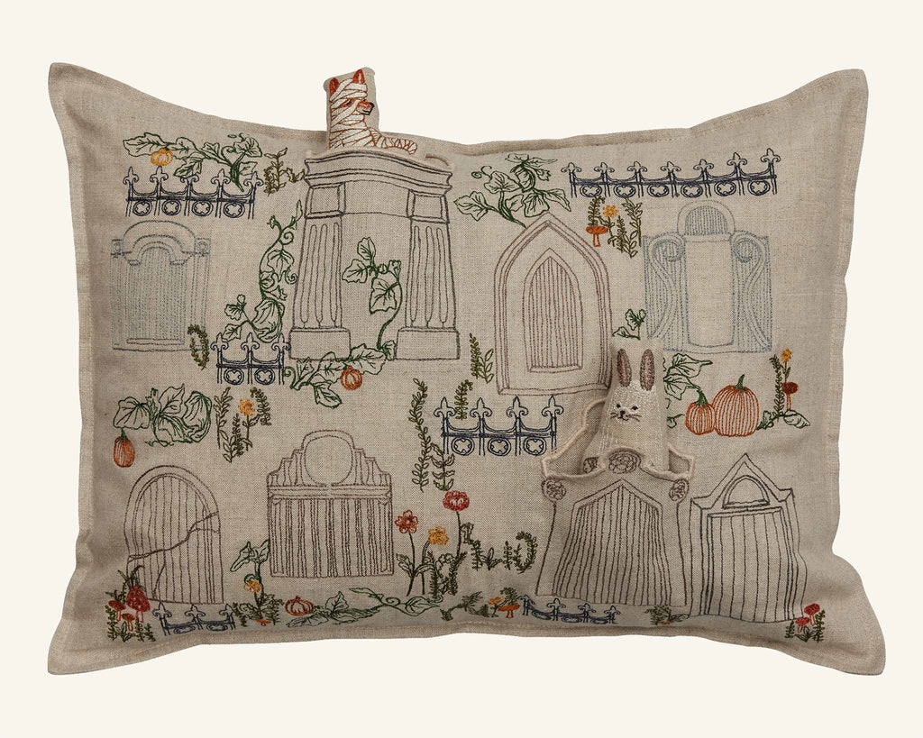 The Coral & Tusk Graveyard Pocket Pillow (ships in one week) is beige and features embroidered gravestones, pumpkins, vines, flowers, fences, and a gray rabbit peeking from a tomb with a "GRIM GRINNING" scroll.