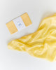 Sarah's Silks Solid Color Playsilk - Sunshine is a folded yellow cloth with a white label next to a shiny, loosely spread playsilk on a white surface—perfect for open-ended play and inspired by Waldorf toys.