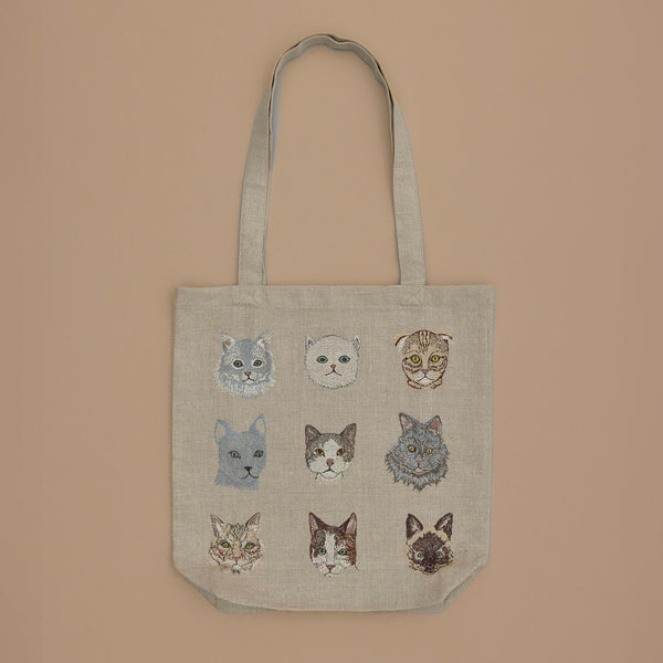 The Cats Tote Bag showcases nine unique cat faces in a 3x3 grid on a beige background, featuring different expressions and fur patterns. It also includes a convenient inside pocket.