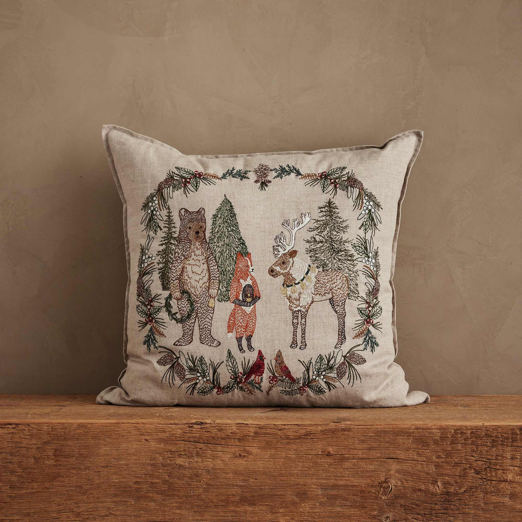 Coral & Tusk Christmas Cheer Wreath Pillow, a square embroidered linen pillow featuring a bear, a fox, and a reindeer standing inside a Christmas wreath decorated with pinecones and cardinals.