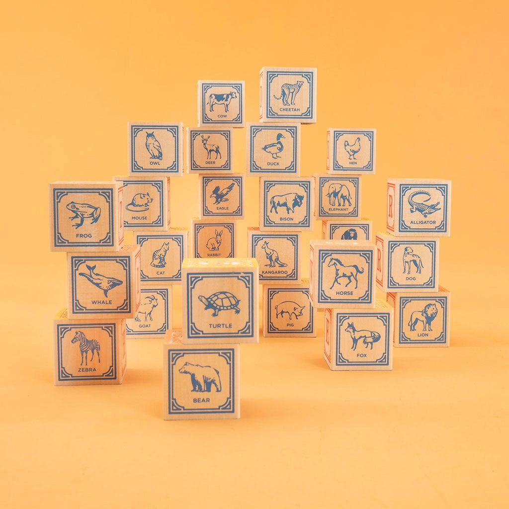 A pyramid of Uncle Goose Classic ABC Blocks with animal illustrations and names, against an orange background. Each block displays a different animal like a frog, whale, or lion in a simple line drawing style.
