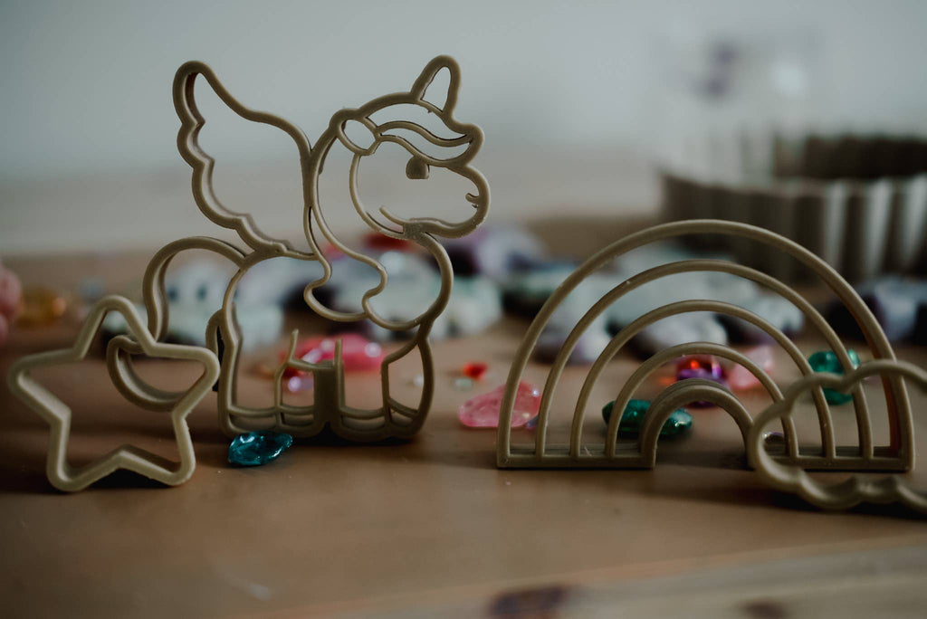 The Unicorn Cutter Set - Eco Playdough Tool includes plastic cookie cutters shaped like a unicorn, a rainbow, and a star, arranged on a table with colorful beads scattered in the background for added sensory play. These eco-friendly tools make creative activities even more magical—especially the enchanting unicorn cutter for imaginative fun.