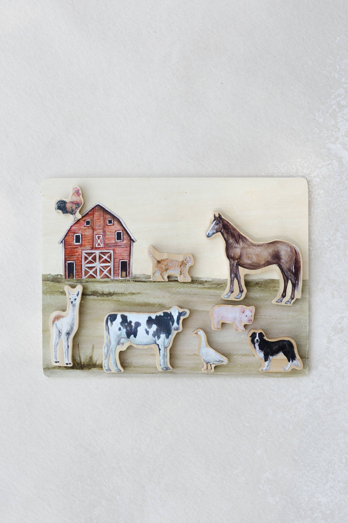 The Farm Animals Wooden Puzzle: A Tabitha Paige Puzzle showcases a red barn scene with a variety of farm animals—including a cow, horse, pig, sheep, dog, rooster, duck, and llama—set on a light background, making it an ideal educational toy for toddlers to learn about their favorite animals.