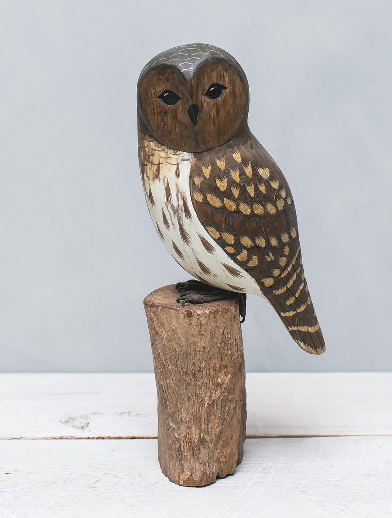 The Hand Carved Ornament - Barred Owl - 13"H features a detailed handmade wooden owl with brown and white feathers, perched on a carved stump against a light gray background and white wooden surface.