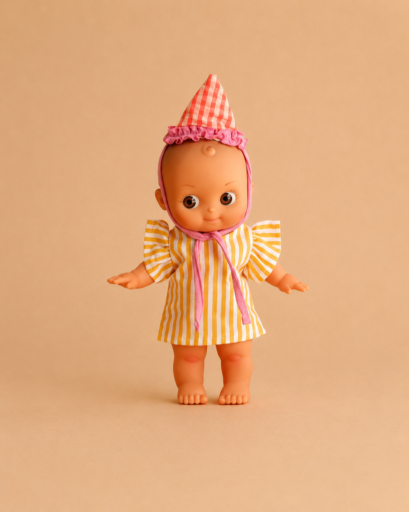 The Minikane Mini Pancha Doll - Ninon with Yellow Striped Birthday Outfit features big eyes and stands barefoot against a plain beige background. She wears a cheerful yellow striped dress and a pink checkered hat, all crafted from high-quality vinyl—making her the perfect companion for imaginative play.
