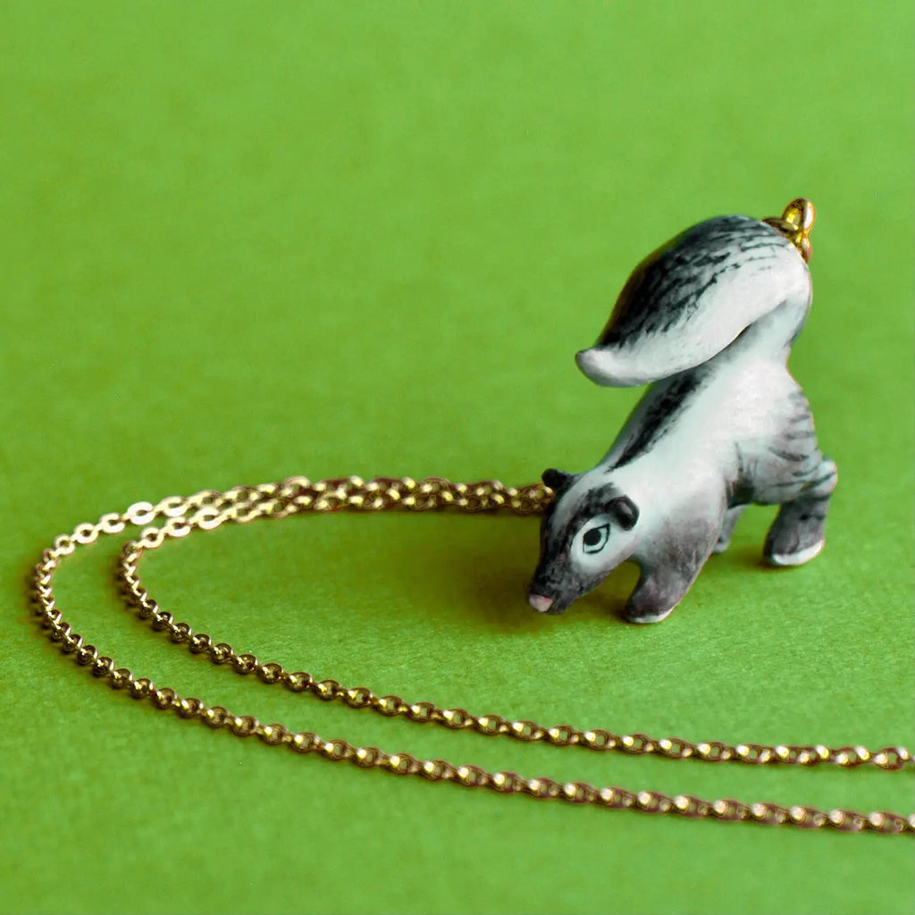 The Skunk Necklace is a hand-painted ceramic pendant shaped like a skunk, with a 24k gold-plated steel chain attached, positioned on a green background. The skunk is predominantly white with grey stripes.