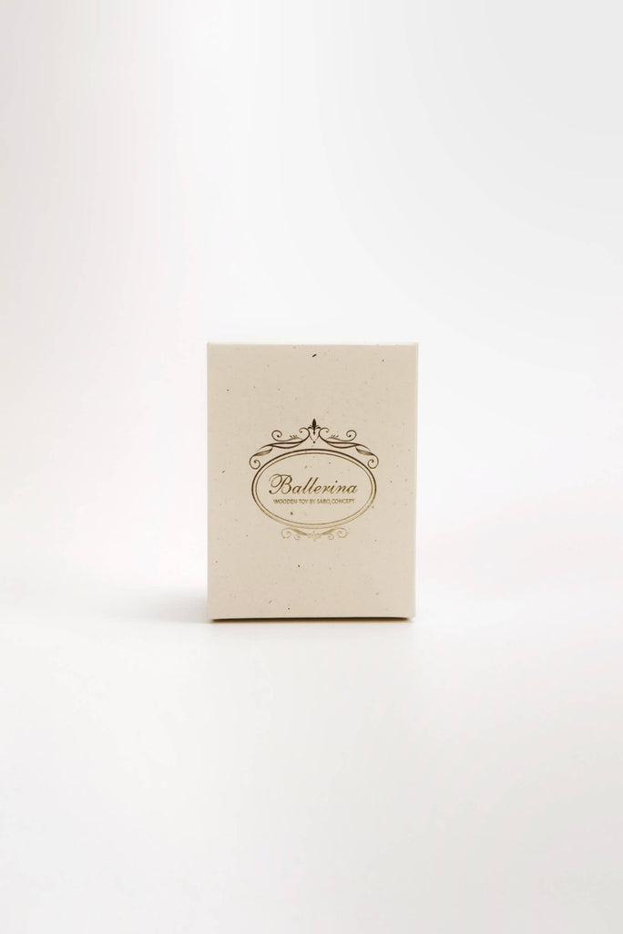 A small beige box labeled "Wooden Ballerina" with an ornate design rests on a white surface, unveiling a delicately hand-painted ballerina figure inside. The box maintains a minimalist yet classy appearance.