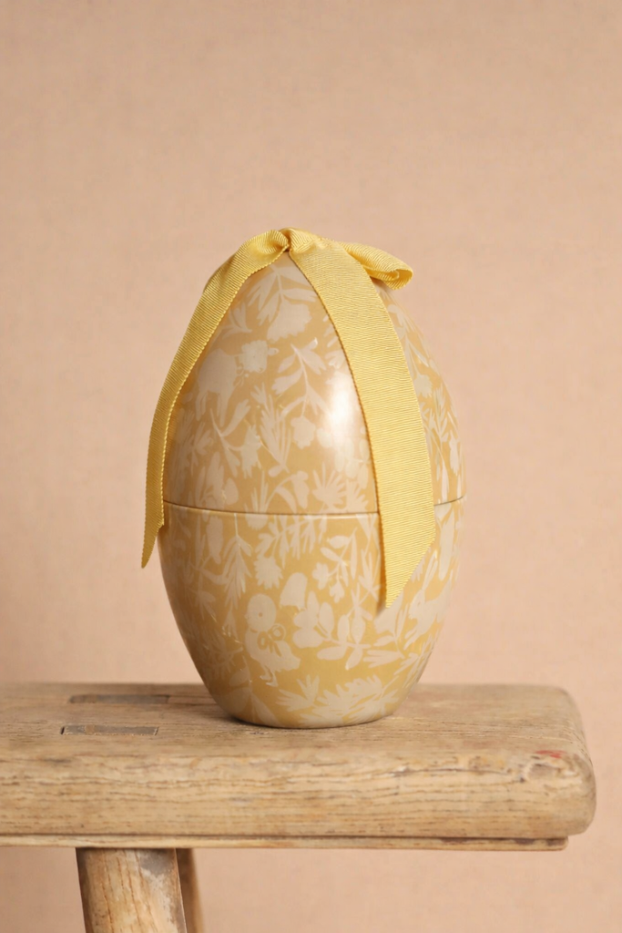 The Easter Basket Gift Set, a pastel yellow decorative egg with floral patterns and a yellow ribbon bow, is displayed on a rustic wooden stool against a beige background.