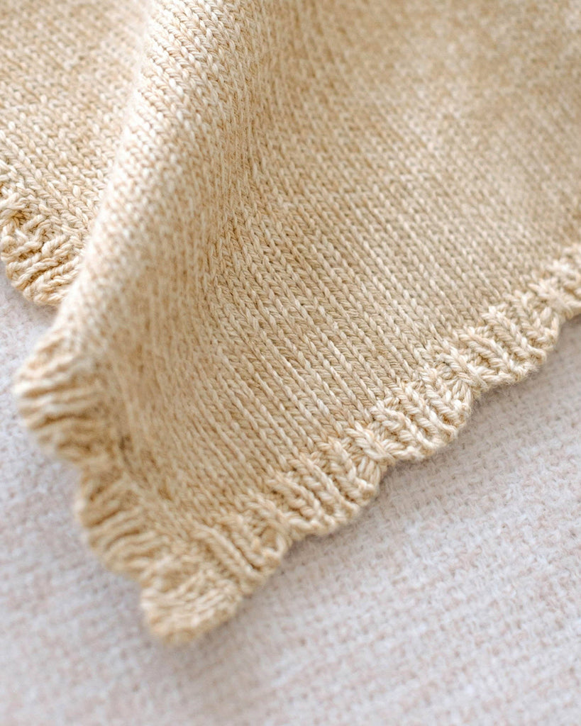 A close-up of the Cuddle + Kind Organic Cotton Baby Bear Lovey in Honey, showing its soft knit texture and scalloped edges, displayed on a light-colored surface with detailed stitches clearly visible.