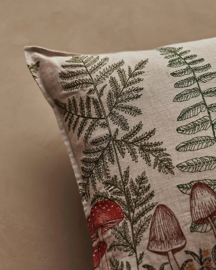A close-up of the Coral & Tusk Mushrooms and Ferns Lumbar Pillow, featuring green ferns and red mushrooms on a beige fabric background.