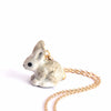 The Goldentail Rabbit Necklace showcases a small, delicate hand-painted porcelain bunny pendant with a speckled gray and white design. The bunny, in a curled position, features a golden tail. The heirloom quality piece hangs on a partially coiled golden chain against a plain white background.