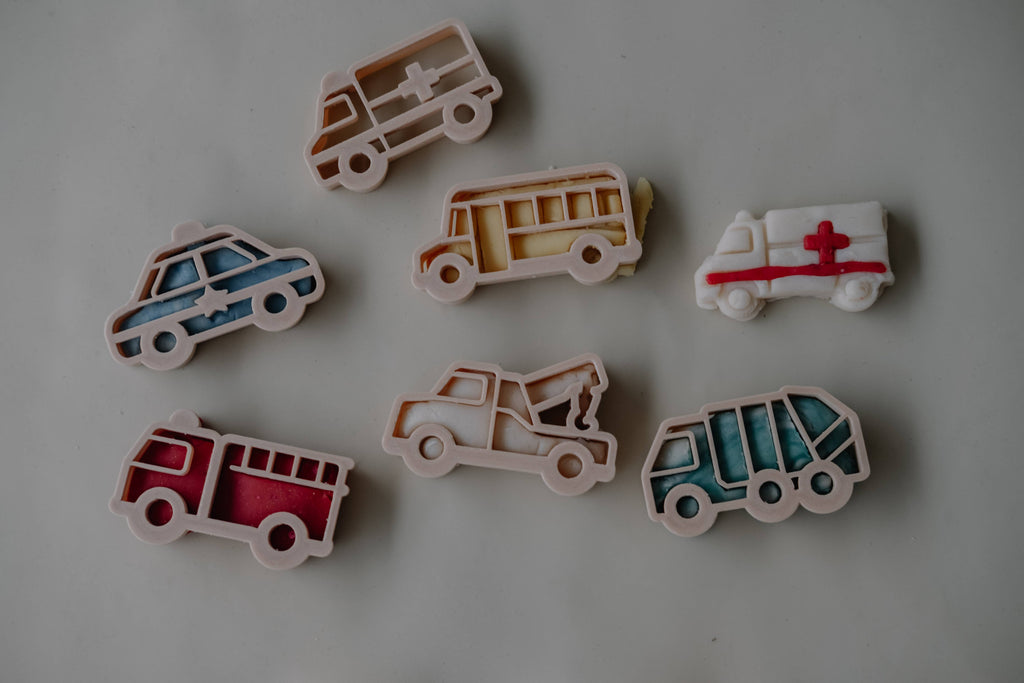 A photo of seven colorful service vehicle cookie cutters from the Service Vehicle Cutter Set - Eco Playdough Tool displayed on a white surface, featuring shapes like a police car, fire truck, ambulance, school bus, tow truck, and two more emergency vehicles. This transport-themed set is perfect for eco playdough activities and sensory play.
