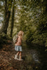 A young child with long blonde hair, pink sweater, denim skirt, and rain boots stands by a muddy creek in a lush forest, carrying the Children's Backpack - Turtle with adjustable shoulder straps.