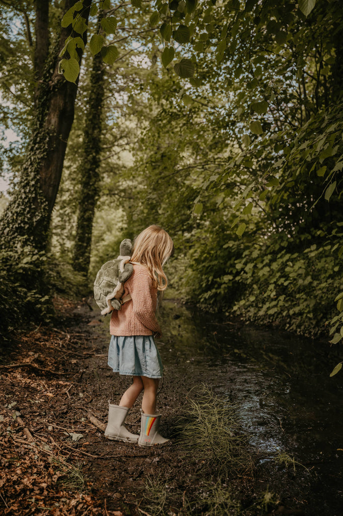 A young child with long blonde hair, pink sweater, denim skirt, and rain boots stands by a muddy creek in a lush forest, carrying the Children's Backpack - Turtle with adjustable shoulder straps.