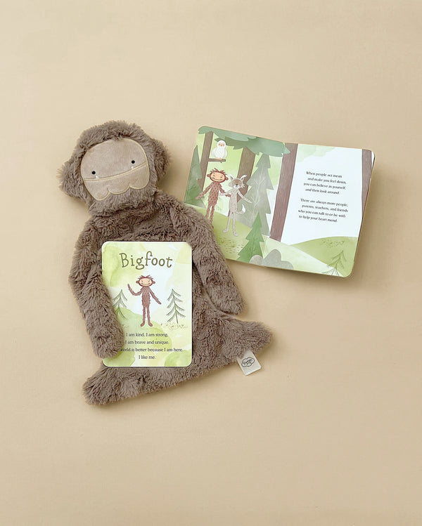 A plush Slumberkins Bigfoot snuggler with a neutral expression is lying next to an open children's book about bigfoot, aimed at boosting children's self-confidence. The book and toy are on a soft.