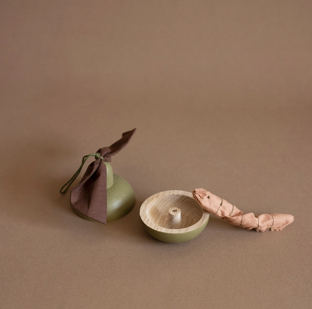 The Wooden Pear & Little Friend, a small, green gourd-shaped container adorned with a brown cloth ribbon at the top, is partially open, revealing a coiled ginger root inside. The eco-friendly toy sits gracefully against a plain, light brown background.