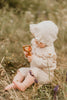 A baby in a bonnet and patterned outfit sits on dry grass, holding the Sabo Concept Bear and Rings Rattle - Yellow and Pink. The wooden rattle supports motor skills, with blurred wildflowers and greenery forming a soft outdoor backdrop.