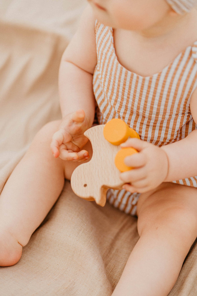 A baby in a striped outfit sits on beige fabric, holding the Sabo Concept Rolling Duck - Yellow toy. The baby's face is partially out of the frame.