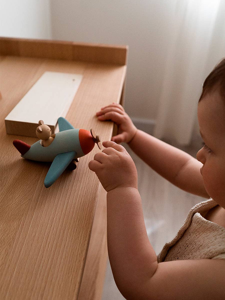 A toddler stands by a wooden table, engaging in imaginative play with the Sabo Concept Aviator Mouse toy airplane.