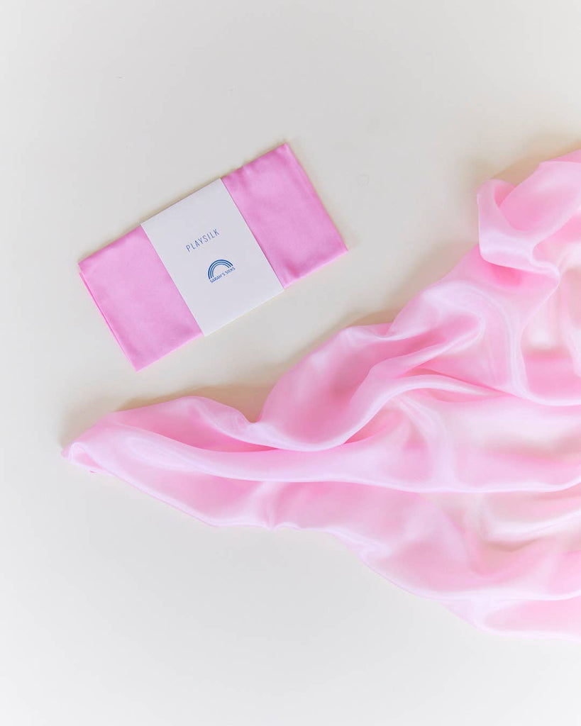 A folded pink Sarah's Silks Solid Color Playsilk sits next to a larger, flowing piece of the same playsilk in matching pink—both ideal for imaginative, open-ended play on a white surface.