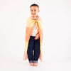 A young child smiles while standing barefoot, reveling in imaginative play. They wear jeans, a white shirt, and a colorful cape made from Sarah's Silk Enchanted Playsilk - Desert in shades of orange and yellow. The plain white background highlights their playful spirit.