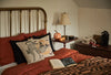 A cozy bedroom features a brass bed, orange bedding, and the Coral & Tusk Supermoon Spell Pillow with a witchy fox and black cat design. An open book sits on the bed; the nightstand holds a lamp, pumpkin, and autumnal decor.