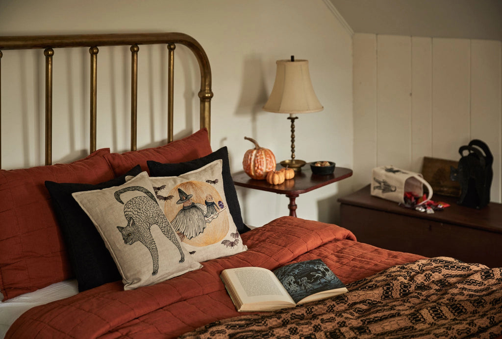 A cozy bedroom features a brass bed, orange bedding, and the Coral & Tusk Supermoon Spell Pillow with a witchy fox and black cat design. An open book sits on the bed; the nightstand holds a lamp, pumpkin, and autumnal decor.