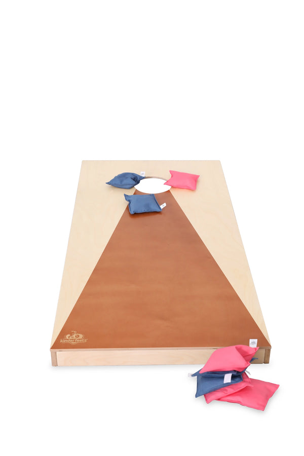 A regulation-sized cornhole board with a triangular brown surface leading to a circular hole near the top is featured in this Cornhole Game. Several blue and pink all-weather bean bags are scattered on the board and on the ground beside it, set against a plain white background.