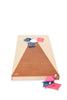 A regulation-sized cornhole board with a triangular brown surface leading to a circular hole near the top is featured in this Cornhole Game. Several blue and pink all-weather bean bags are scattered on the board and on the ground beside it, set against a plain white background.