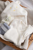 A wicker basket contains a baby hooded garment with a cute mouse face and ears, a pair of baby socks (one white, one blue), and a small white flower. A Baby Swaddle is also included. An organic cotton baby swaddle suitable for 0-3 months completes the set, all neatly arranged on soft fabric.