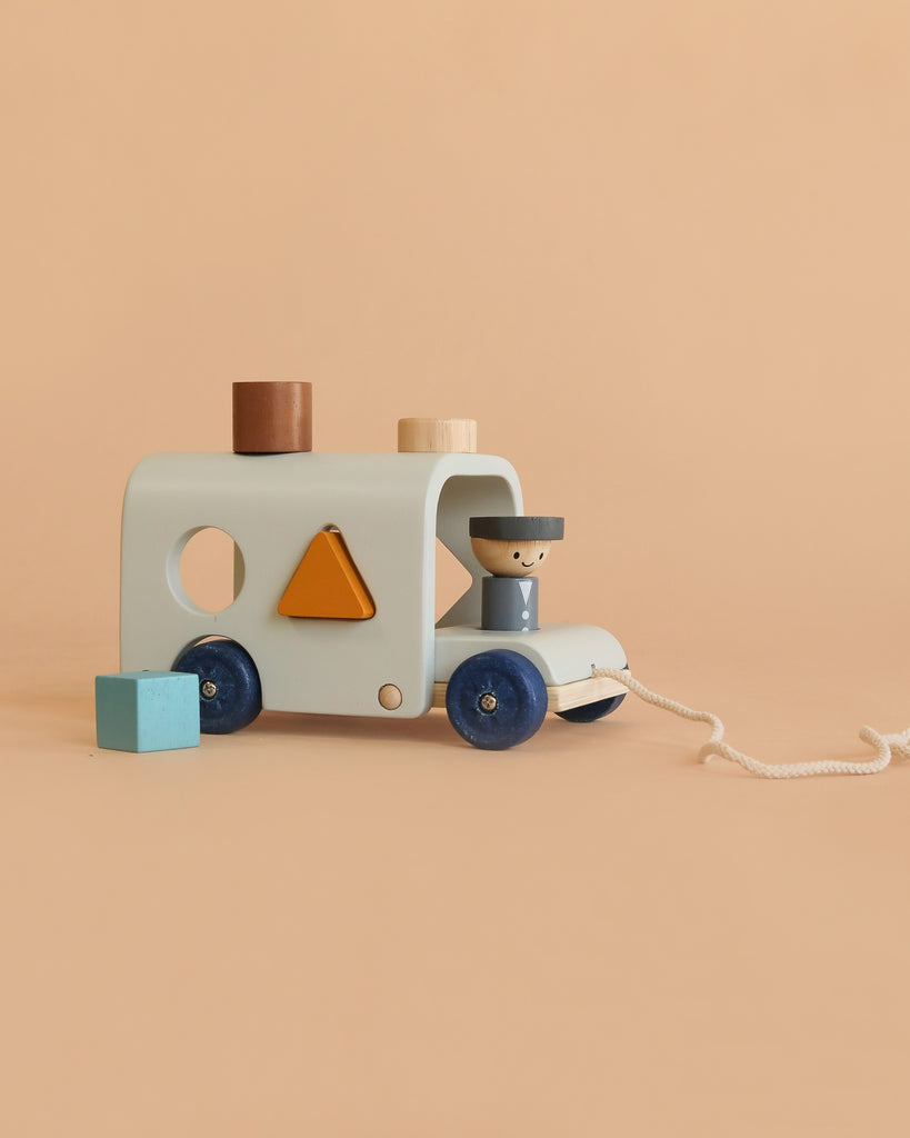 Introducing the Sorting Bus – Orchard Series: This charming wooden toy features shape sorter pieces on top and includes a blue driver figure inside, inspired by the classic PlanToys design. The bus doubles as both a pull toy and a shape sorter, with a white pull string attached to the front. A blue block sits close by, all set against a plain light tan background.