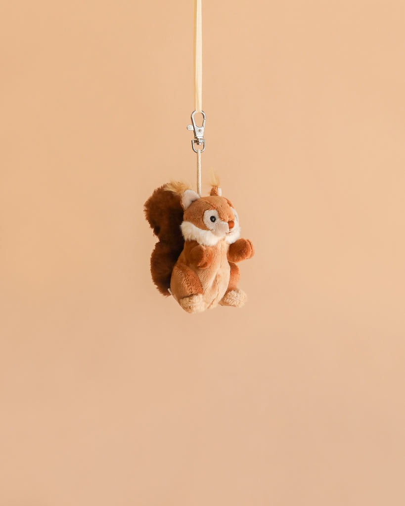 The Bukowski - Brunis Squirrel Bag Charm, a small plush woodland companion, dangles from its beige strap against a simple light brown background.