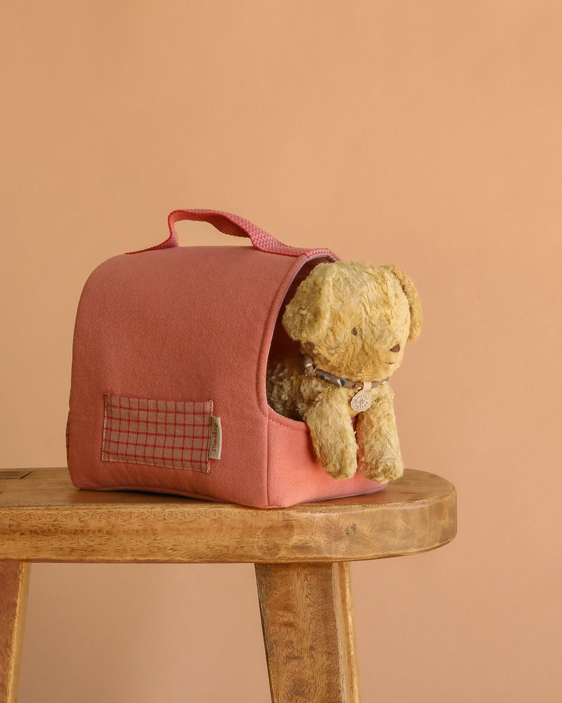 The Maileg Puppy in Coral Pet Carrier - Soft Sand, a small tan plush puppy, sits inside a pink pet carrier on a wooden stool against a plain beige background.