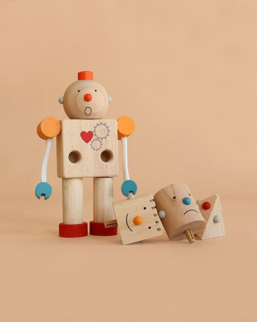 Build -A- Robot, a wooden toy robot with a heart on its chest, stands upright with two interchangeable heads—one smiling and one sad—placed at its feet. Perfect for teaching emotions, the scene is set against a solid beige background.