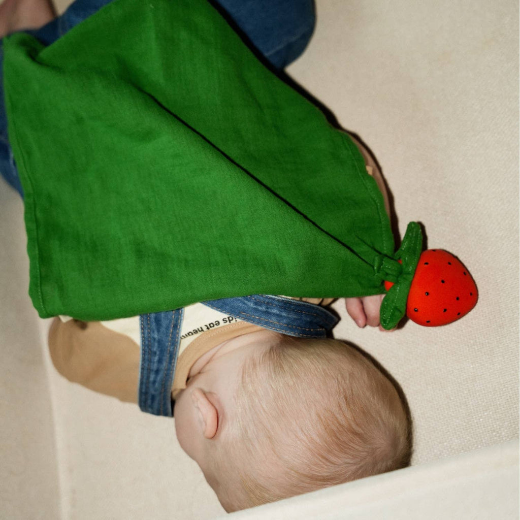 A baby lies on its side, wearing a tan shirt and blue overalls, holding the Sweetie the Strawberry Big Doudou—an organic cotton doudou with a green cloth body and a natural rubber strawberry-shaped head.