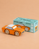 A wooden racing car in orange and white sits in front of its teal box labeled “No. 22 GT125 Cami | Made in England.” The box features illustrations and the words “Limited Edition.” Reminiscent of classic American muscle, this striking toy racing car stands out against the beige background.