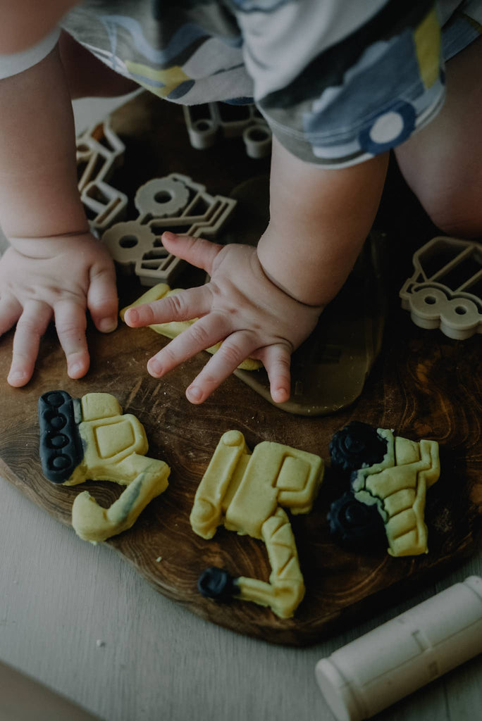 A child presses their hands on a wooden surface surrounded by construction vehicle-shaped cookies and matching cookie cutters, enjoying sensory play with our Construction Cutter Set - Eco Playdough Tool.