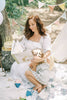 A woman in a white dress reads Winnie-the-Pooh: Finding Feelings to a child on a picnic blanket outdoors, surrounded by party decorations, cake, trees, and bunting.