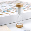 A white hourglass with gold caps stands in the foreground, with a blurred Scrabble Bianco Edition game board in the background.