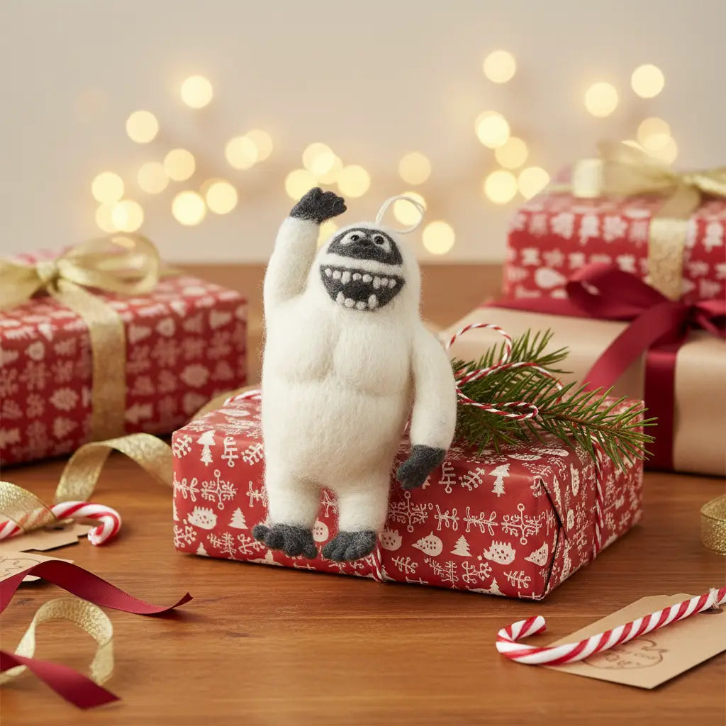 The Handmade Felt Yeti Christmas Tree Ornament, lovingly handcrafted in Nepal, stands on a red gift box amid wrapped presents, candy canes, and glowing holiday lights, smiling brightly for a festive touch.