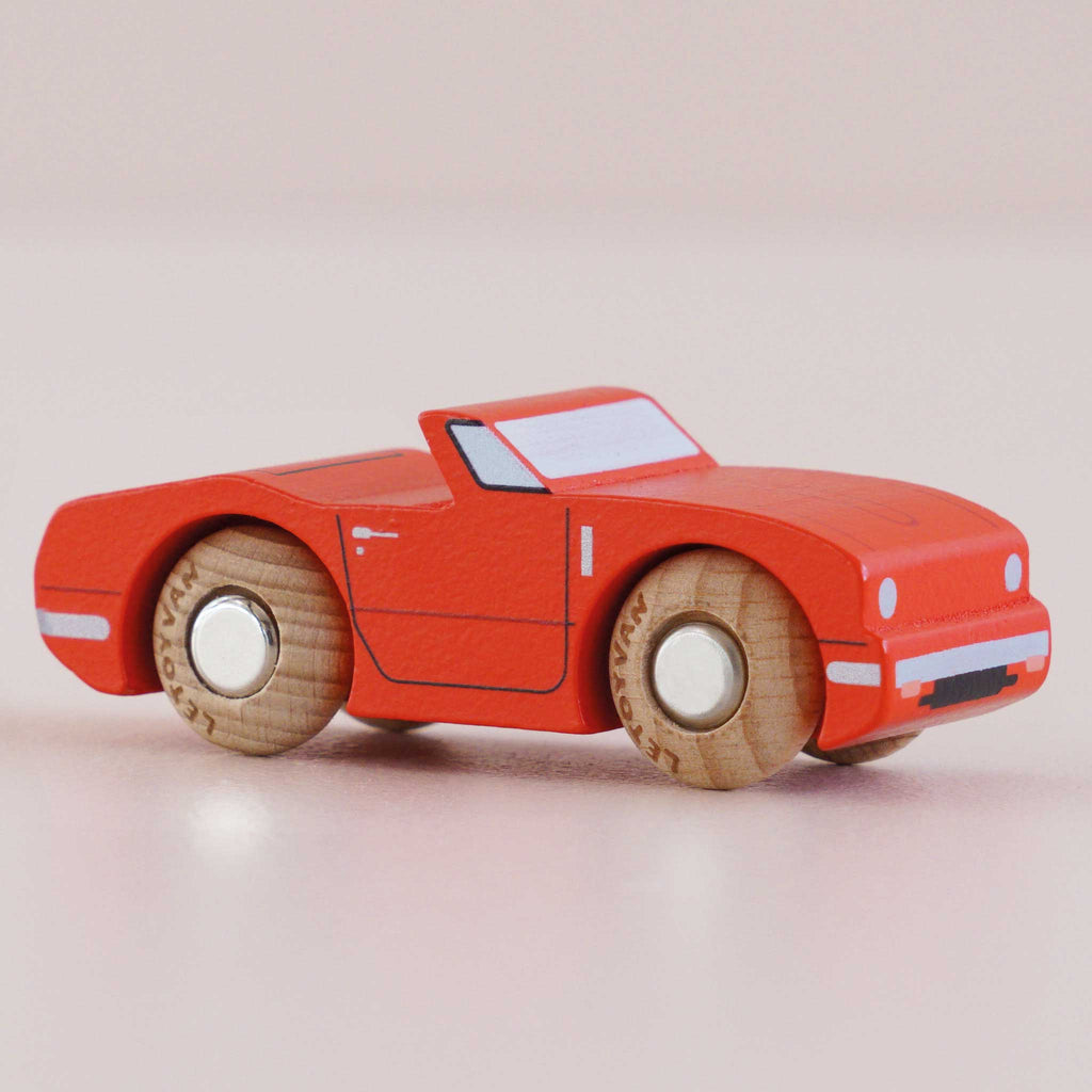 The No. 13 GT75 Marc, made in England, is a classic small wooden convertible car painted in racing red with white accents and natural wooden wheels, pictured on a light pink surface.