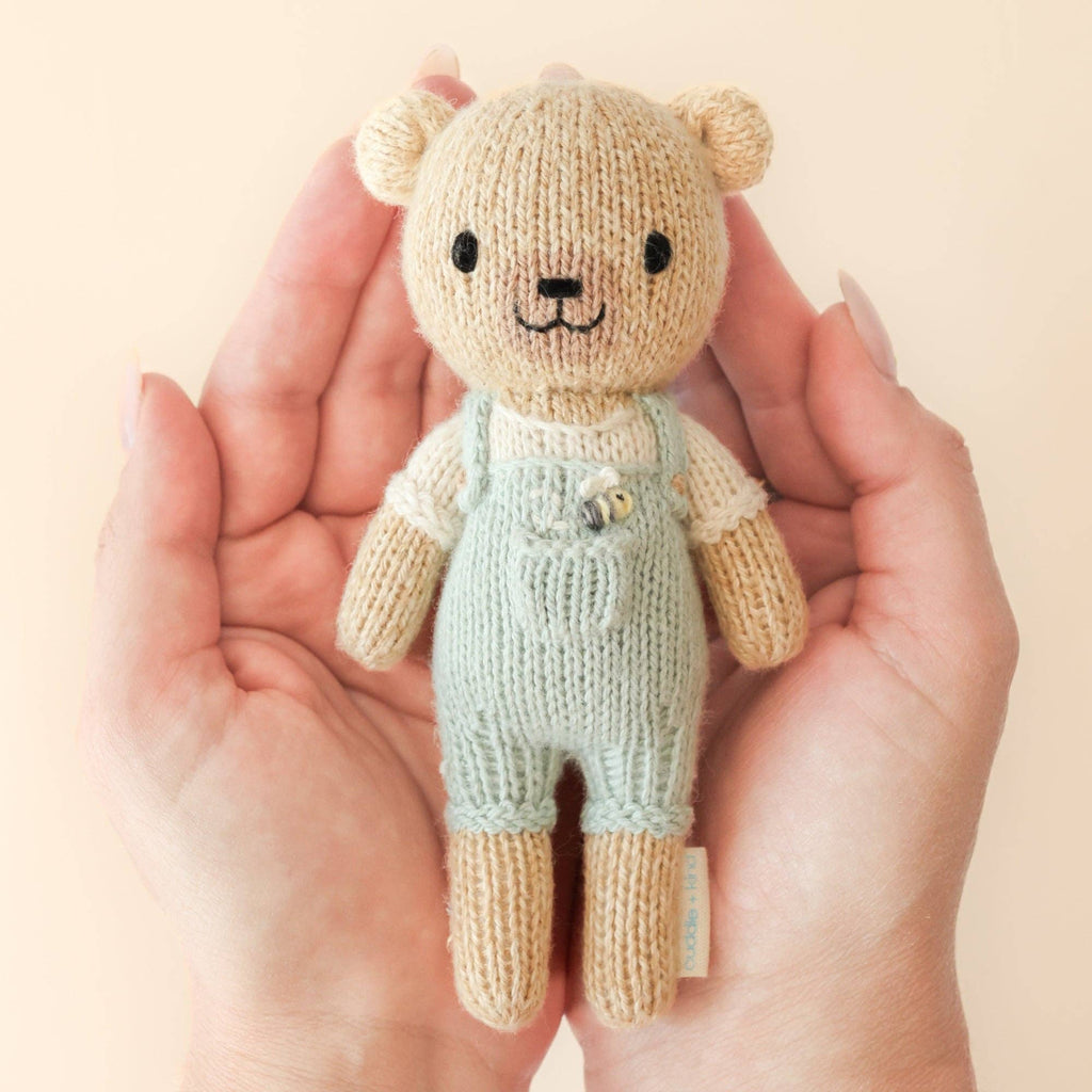 A pair of hands gently holds a Cuddle + Kind Tiny Charlie the Honey Bear, a hand-knit doll in mint green overalls and a cream shirt, against a light beige background.